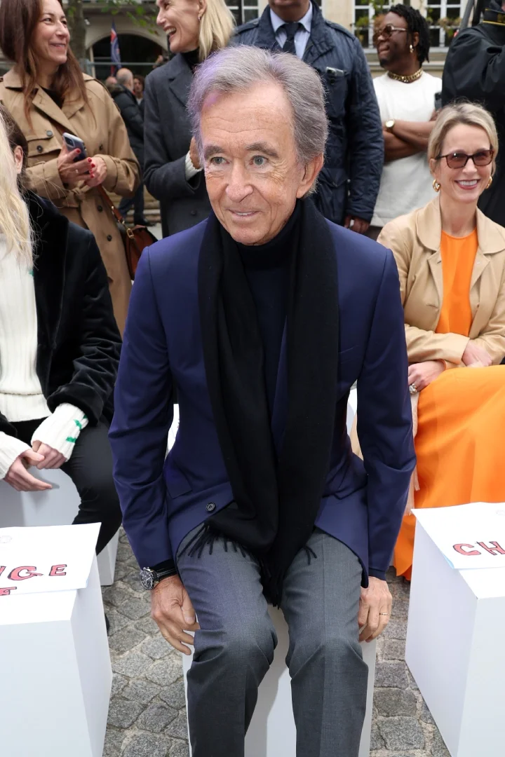 Bernard Aranult sitting showing off his watch during a fashion show,, in a black turtleneck and blue suit jacket