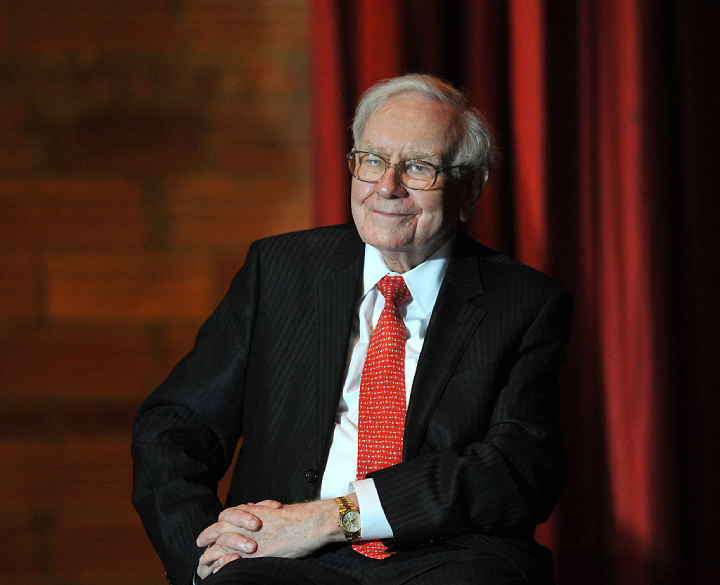 Warren Buffett posing for the camera in a full black suit, with his Rolex President visible on his left wrist
