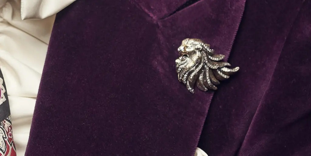 A lion's head brooch in a lapel of a purple velvet jacket. 