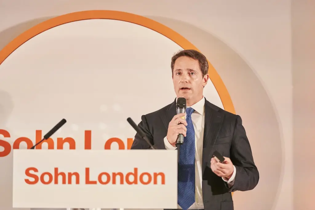 Carson Block wearing a suit and holding a remote control speaking at the Sohn London holding a microphone