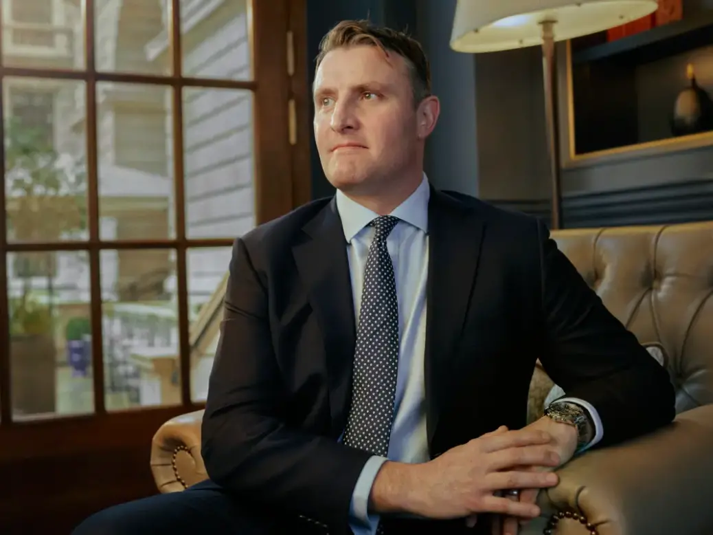 Ollie Barnett wearing a suit and spotted tie on an armchair