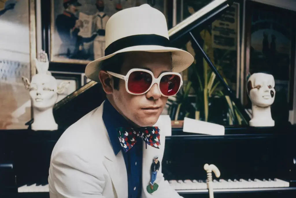 Elton John wearing sunglasses and a hat sitting at a piano