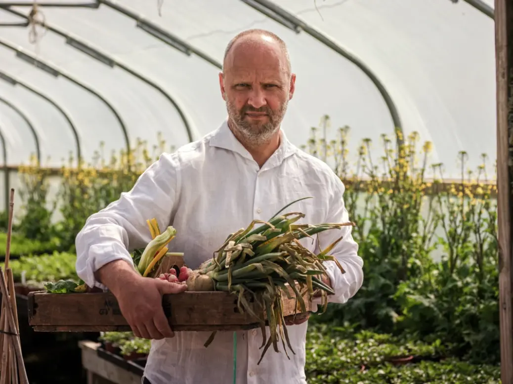 Simon Rogan holding a wooden box with vegetables in it in a greenhouse